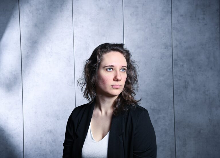 Frau im Alter von 30 mit langen Haaren ist schräg zur Kamera zugewandt. Die Perspektive reicht bis kurz unterhalb ihrer Brust. Sie trägt ein weißes T-Shirt mit schwarzem Blazer. Sie schaut zur Seite, nicht in die Kamera. Der Hintergrund ist eine graue Wand mit eher dunkler Beleuchtung.