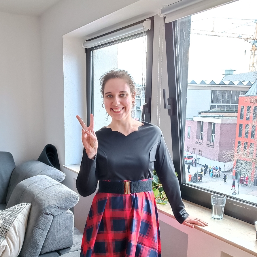 Eine Frau um die 30 steht vor einem Fenster in einem hellen Raum mit Stadtblick und lächelt herzlich in die Kamera. Sie zeigt ein Peace-Zeichen und trägt ein elegantes schwarzes Oberteil mit wellenförmigem Ausschnitt, kombiniert mit einem rot-blau karierten Midirock und einem schwarzen Gürtel mit goldfarbener Schnalle.